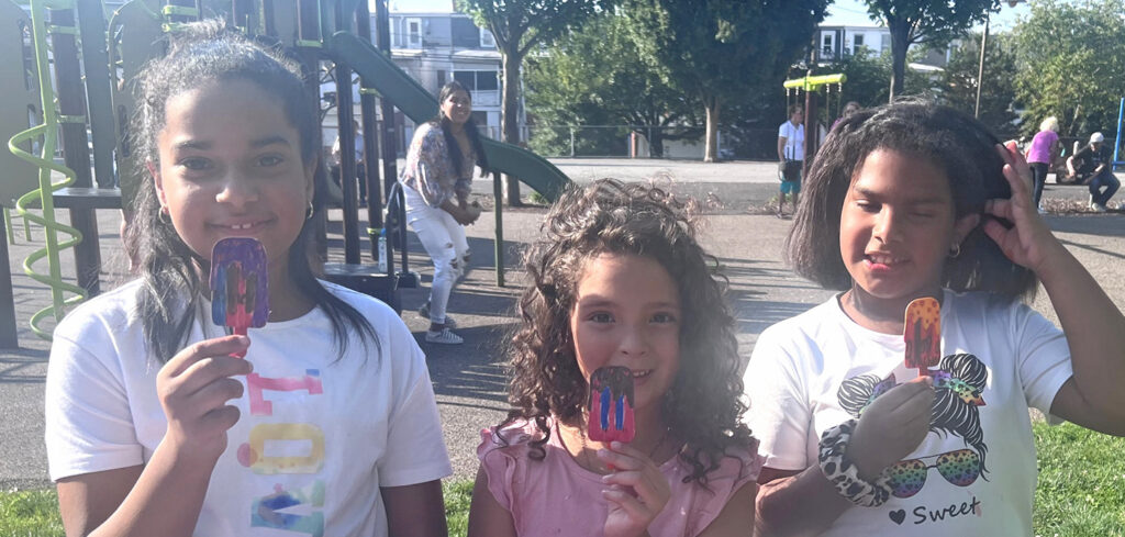 Three young girls at a playground smiling and holding popsicle art they created.